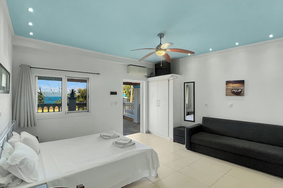 A bedroom with a bed, a sofa, a ceiling fan, and a window with a view of the sea.