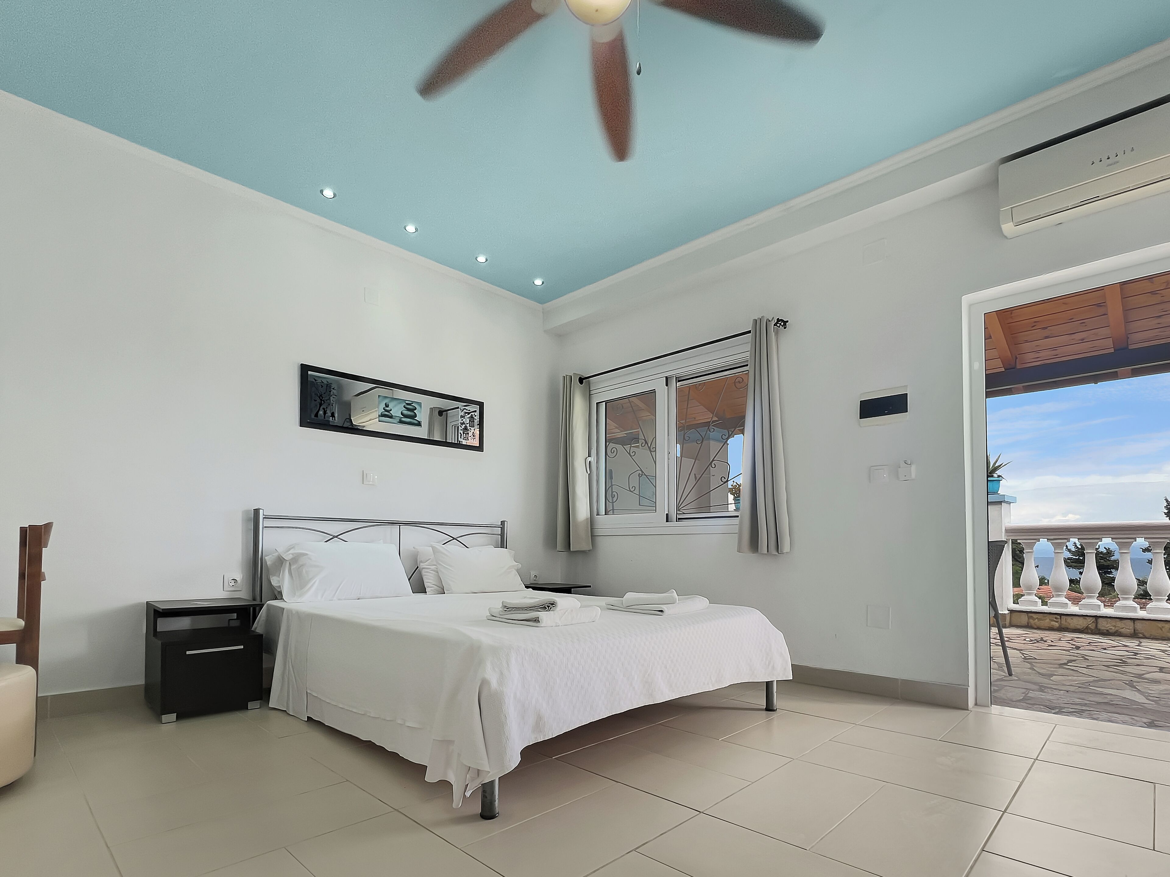 Una camera da letto con un letto, un comodino, una ventola a soffitto, una finestra con tende e vista del balcone esterno.
