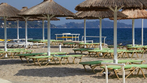 A beach with rows of green lounge chairs and thatched umbrellas, a table, and a view of the sea and distant hills. - Andromaches Holiday Apartments (Corfu)