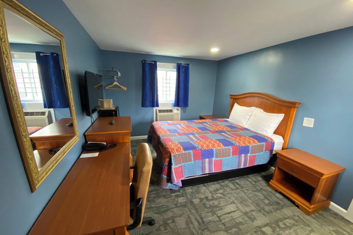 A hotel room with a wooden desk, a bed with a colorful quilt, a nightstand, a mirror, and a window with curtains.