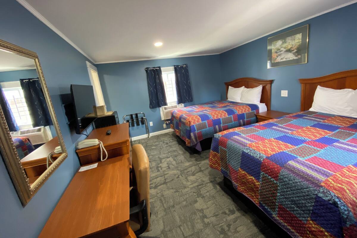 A hotel room with two beds, a desk, a chair, a mirror, a TV, and a window with curtains.