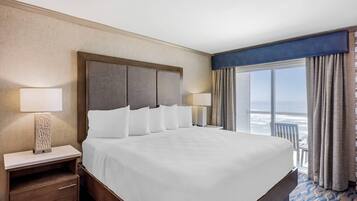 A hotel room with a large bed, two bedside lamps, a nightstand with drawers, and a view of the ocean through a large window with curtains.