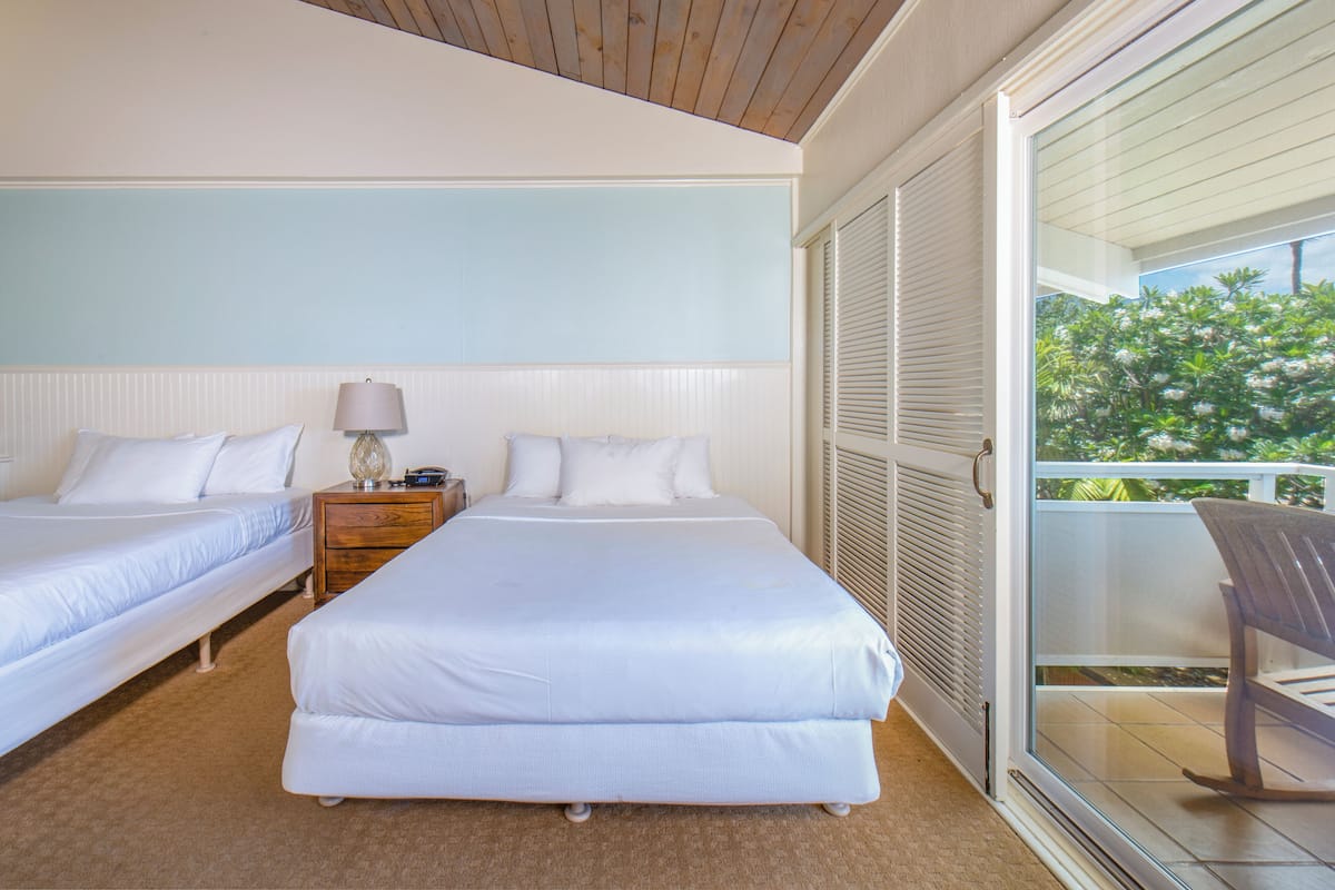 Habitación de hotel con dos camas, una mesita de noche, una lámpara y vistas al exterior.
