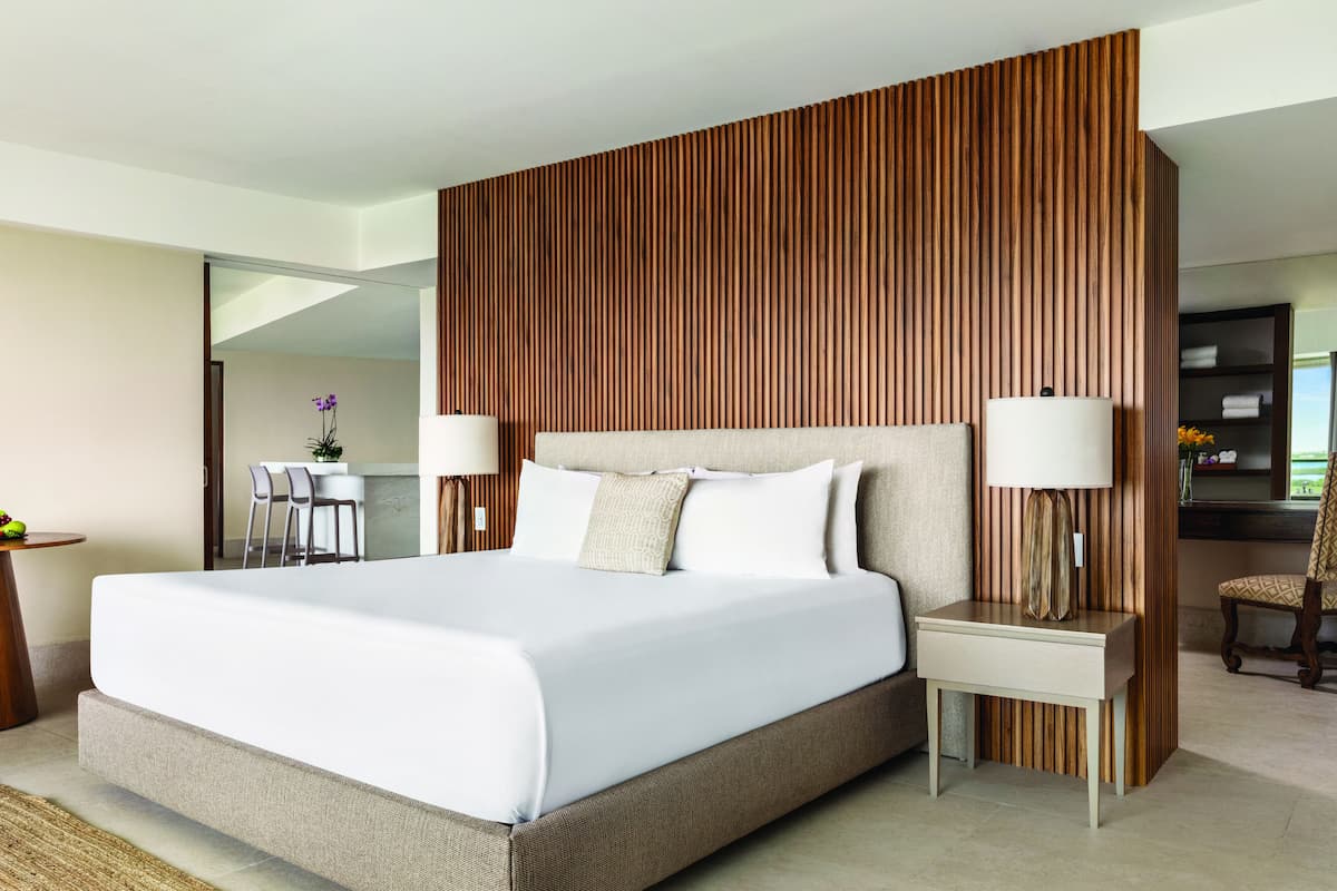A modern hotel room with a large bed, wooden panel wall, two bedside tables with lamps, and a view of the dining area.