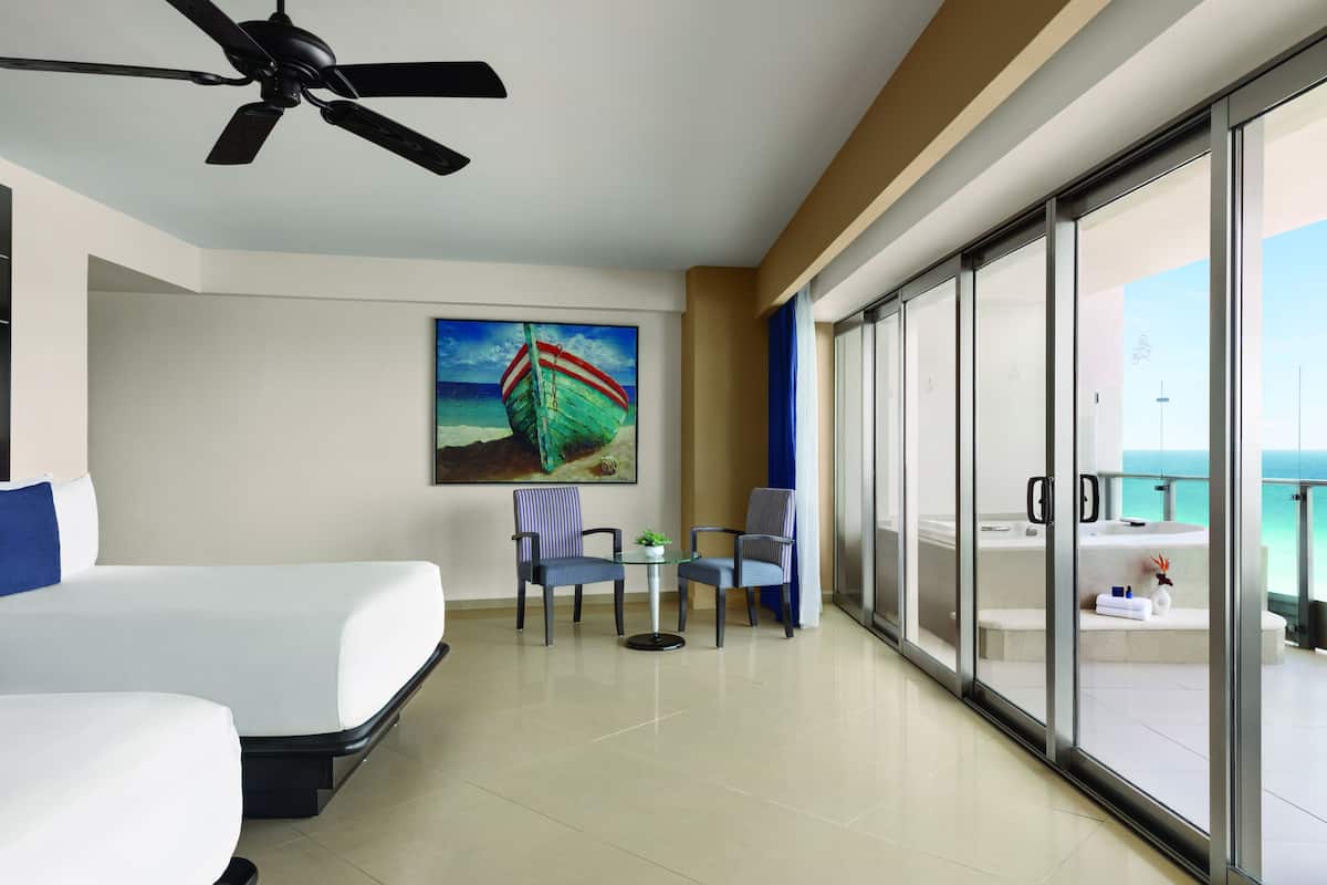 A hotel room with a bed, a ceiling fan, a painting of a boat, a small table with two chairs, and a view of the ocean through large glass doors.