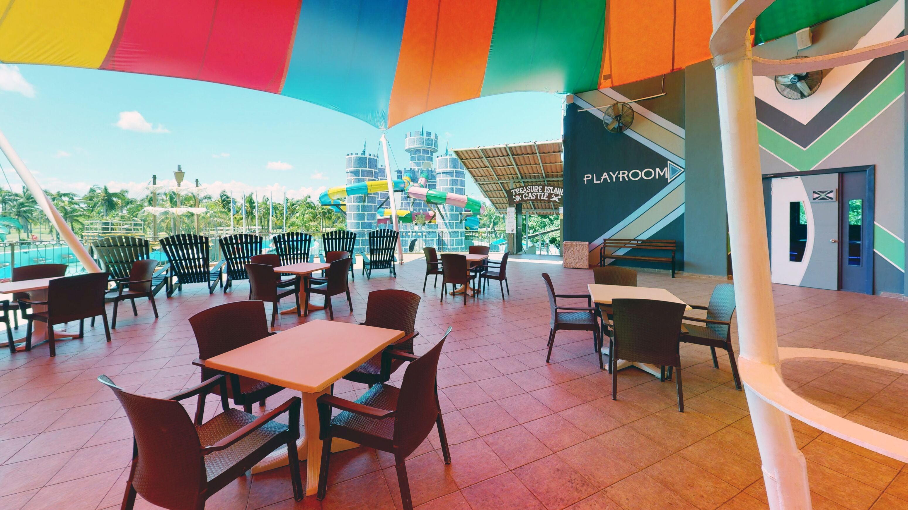 A spacious outdoor dining area with multiple tables and chairs, a play area with slides and play structures, and a sign for 'Playroom'.