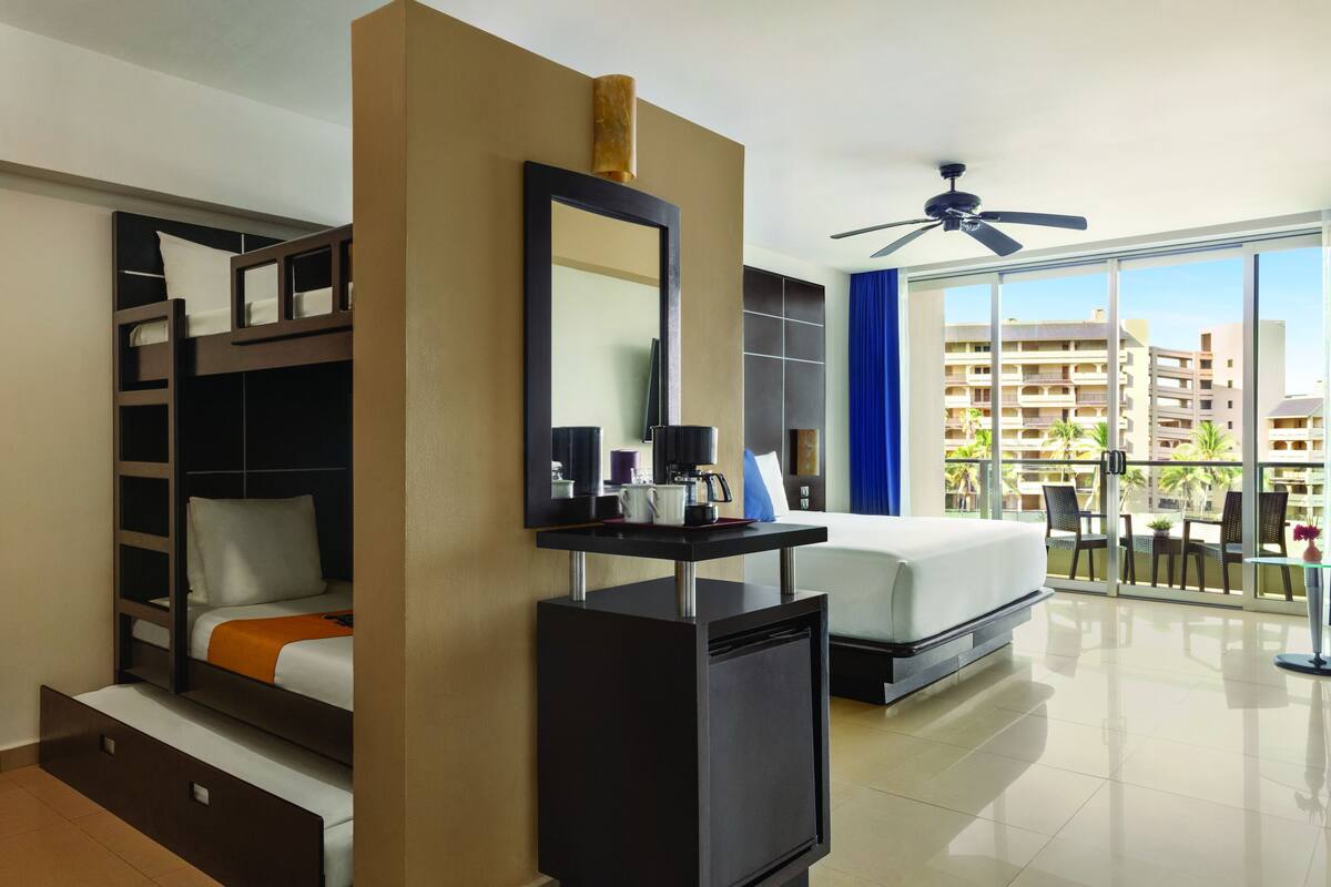 A hotel room with a bunk bed, a desk with a mirror, a ceiling fan, and a balcony with a view of the city.