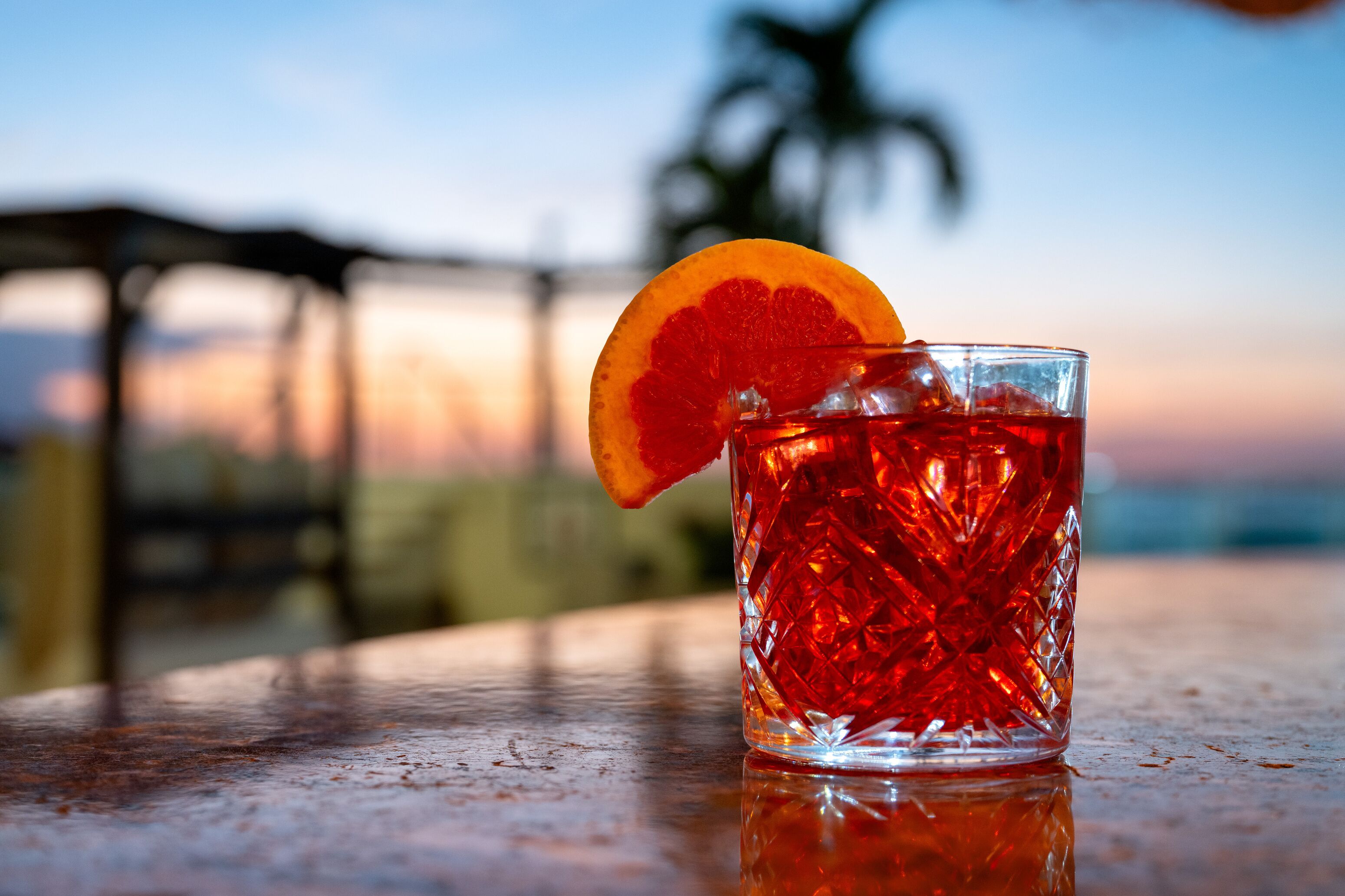 A clear glass with a red drink, a slice of orange garnish, and ice cubes on a table with a blurred outdoor background.