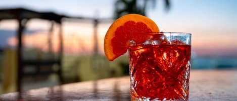 A clear glass with a red drink, a slice of orange garnish, and ice cubes on a table with a blurred outdoor background.