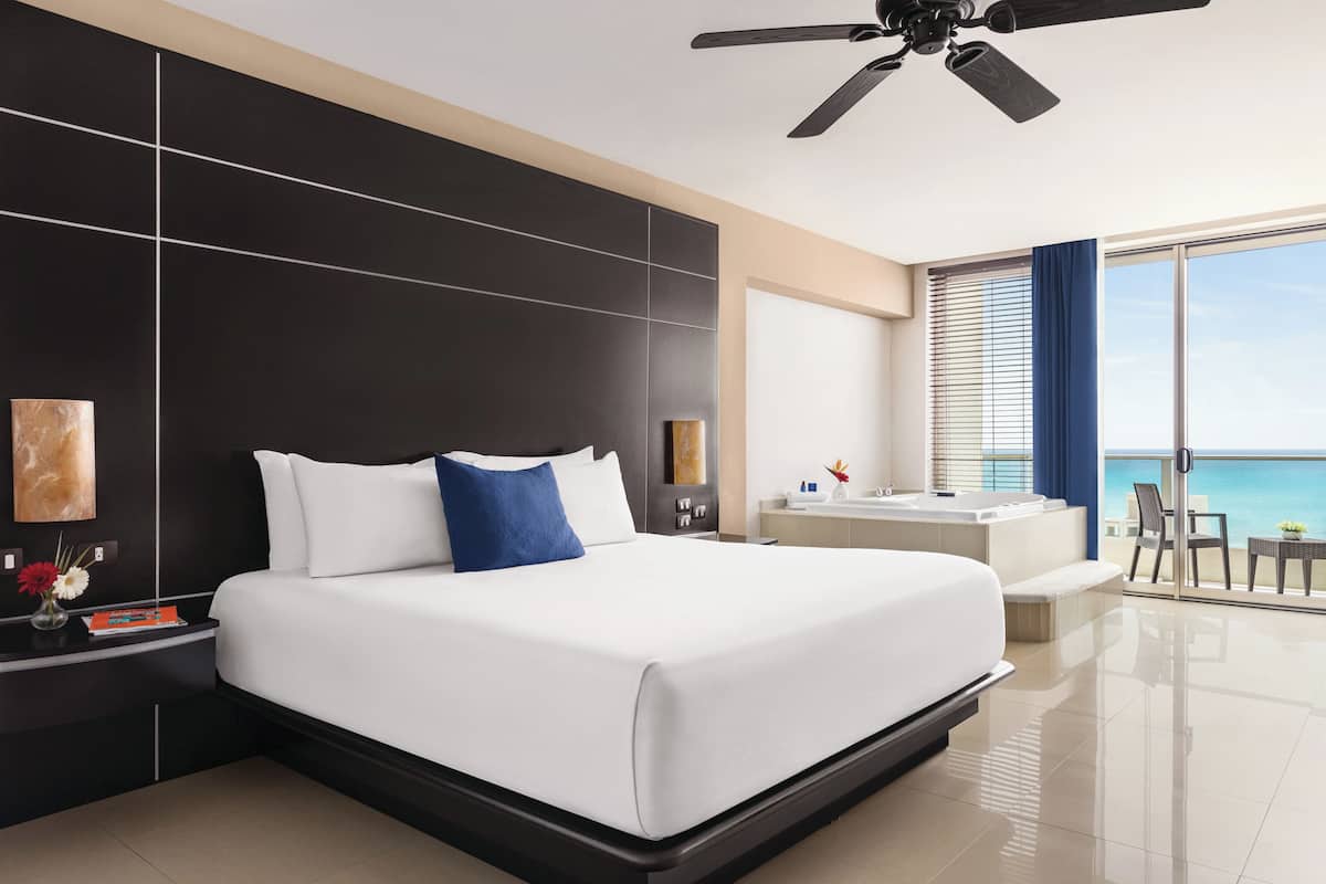 A modern hotel room with a large bed, a ceiling fan, a balcony with a view of the sea, and a separate bathroom area.