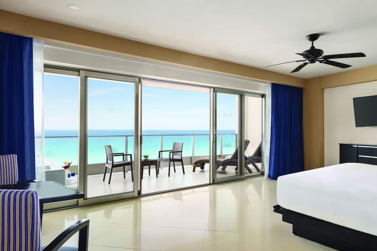 A hotel room with a large bed, a ceiling fan, and a balcony with a view of the ocean.