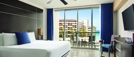 Habitación de hotel con cama, ventilador de techo, televisión y balcón con vistas a edificios y palmeras.