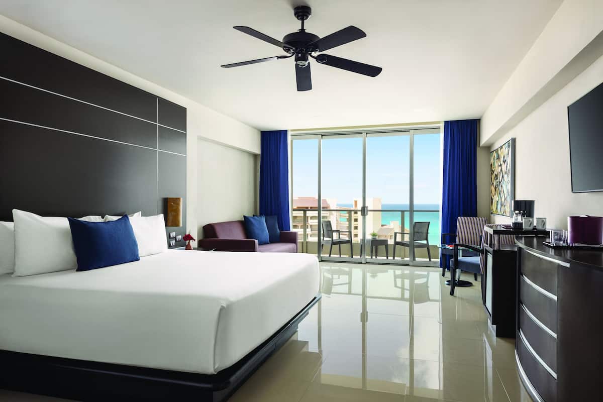 A hotel room with a large bed, a desk, a balcony with a view of the ocean, and a ceiling fan.
