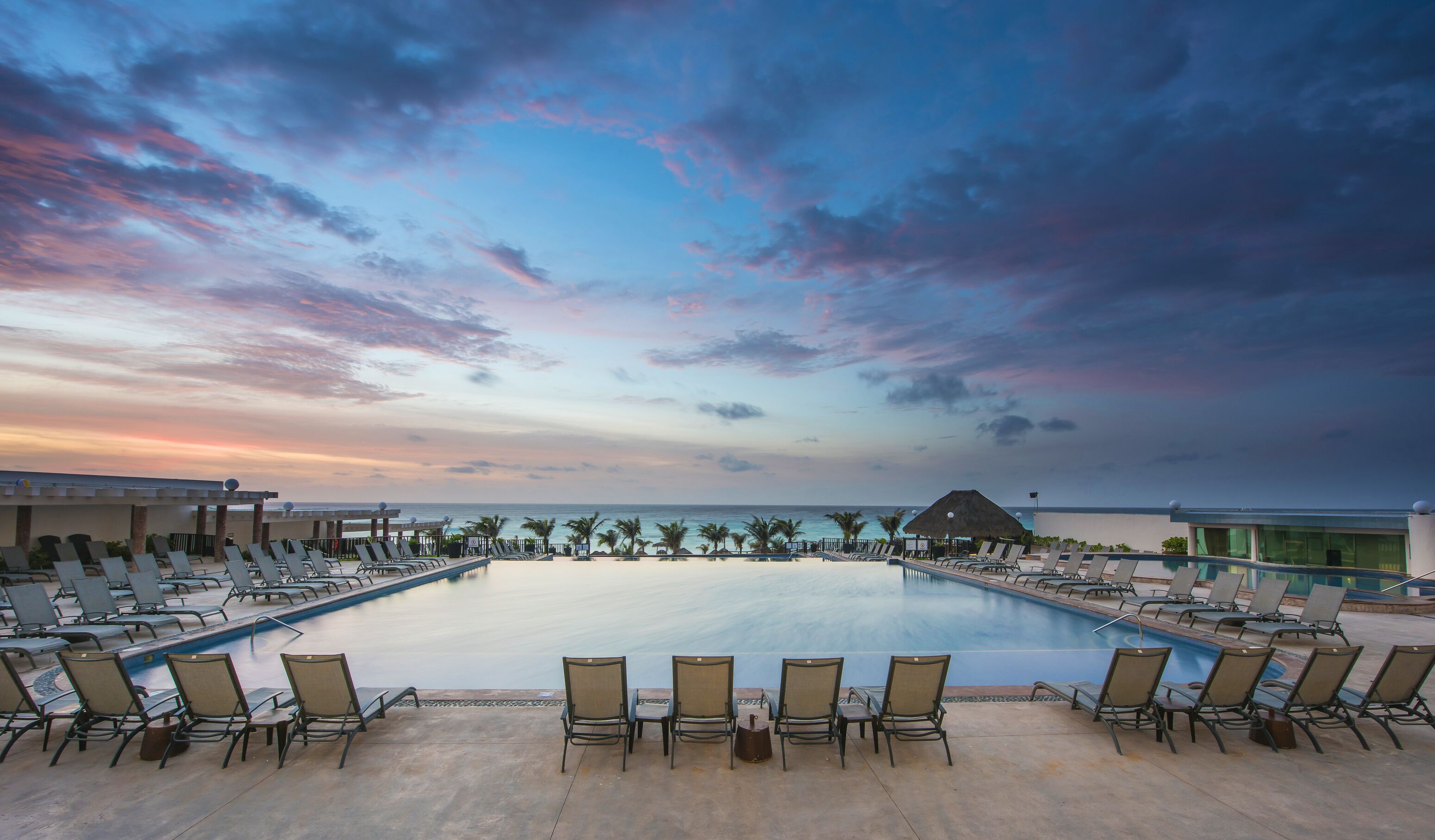 A poolside area with lounge chairs and a view of the ocean at sunset.