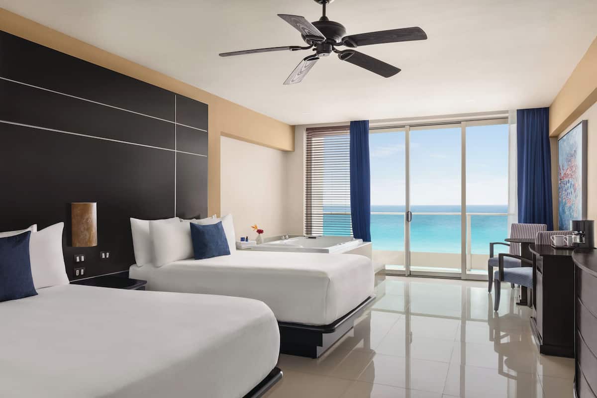 A hotel room with two beds, a desk, a chair, a bathtub, and a view of the ocean.