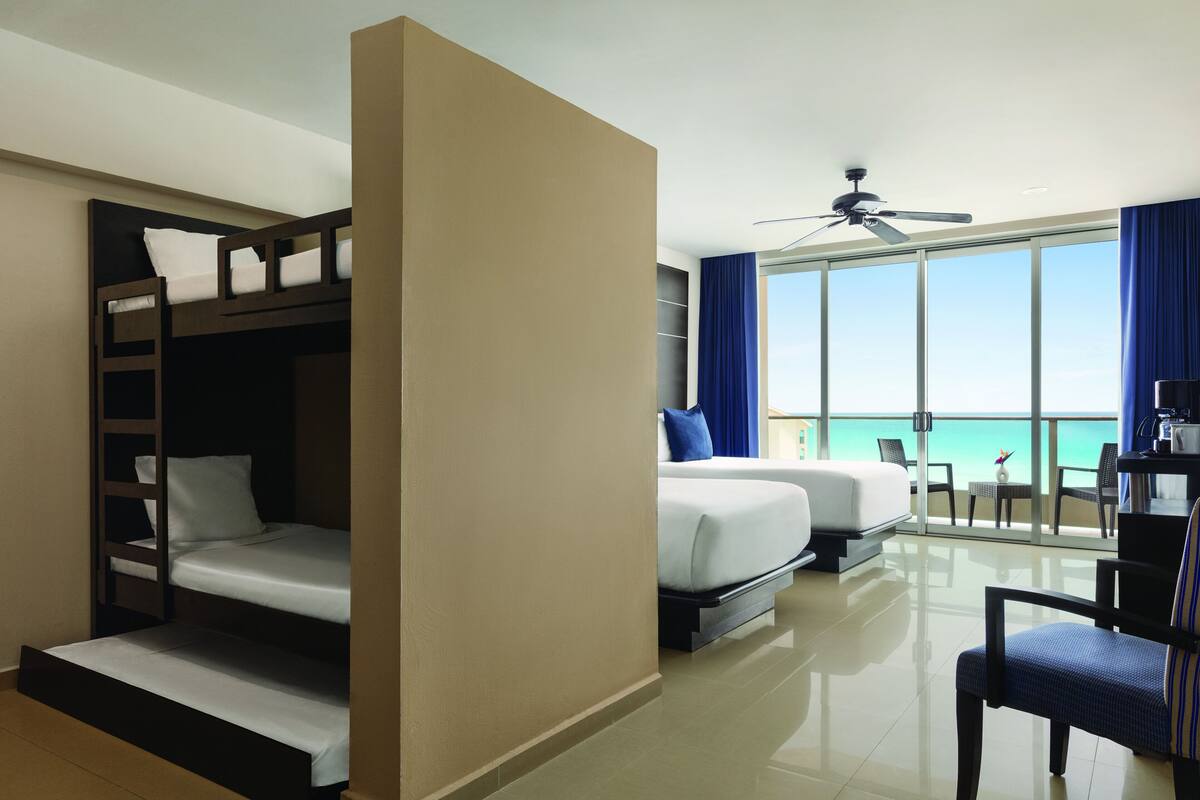 A hotel room with a bunk bed, a separate bed, a balcony with a view of the ocean, a ceiling fan, and a desk with a chair.