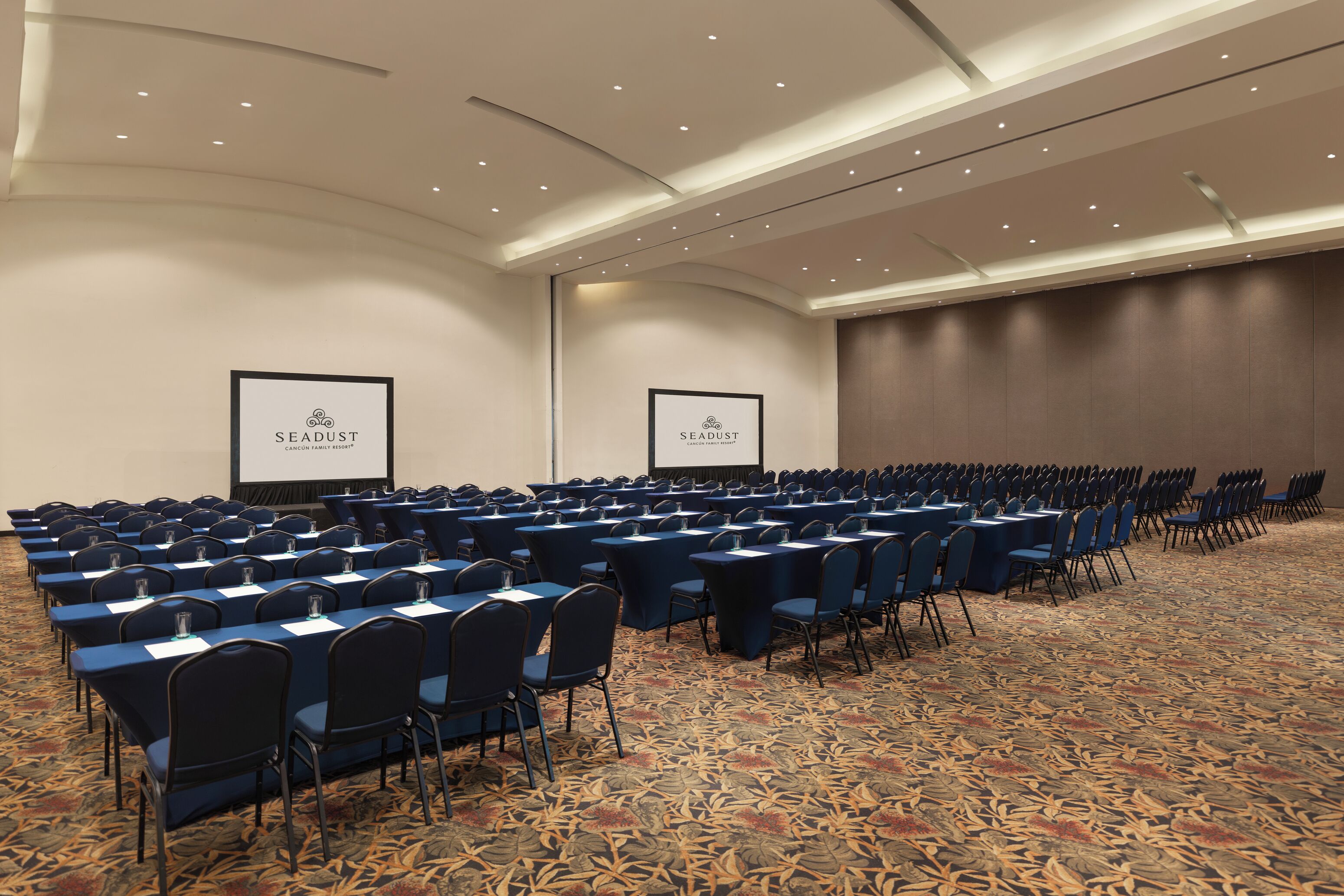 A conference room with rows of tables and chairs, a carpeted floor, and two screens displaying the SEADUST logo.