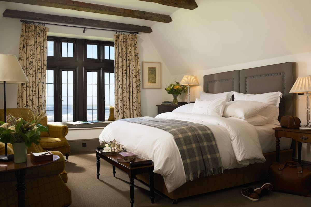A bedroom with a large bed, a window with curtains, a bedside table with a lamp, and a chair.