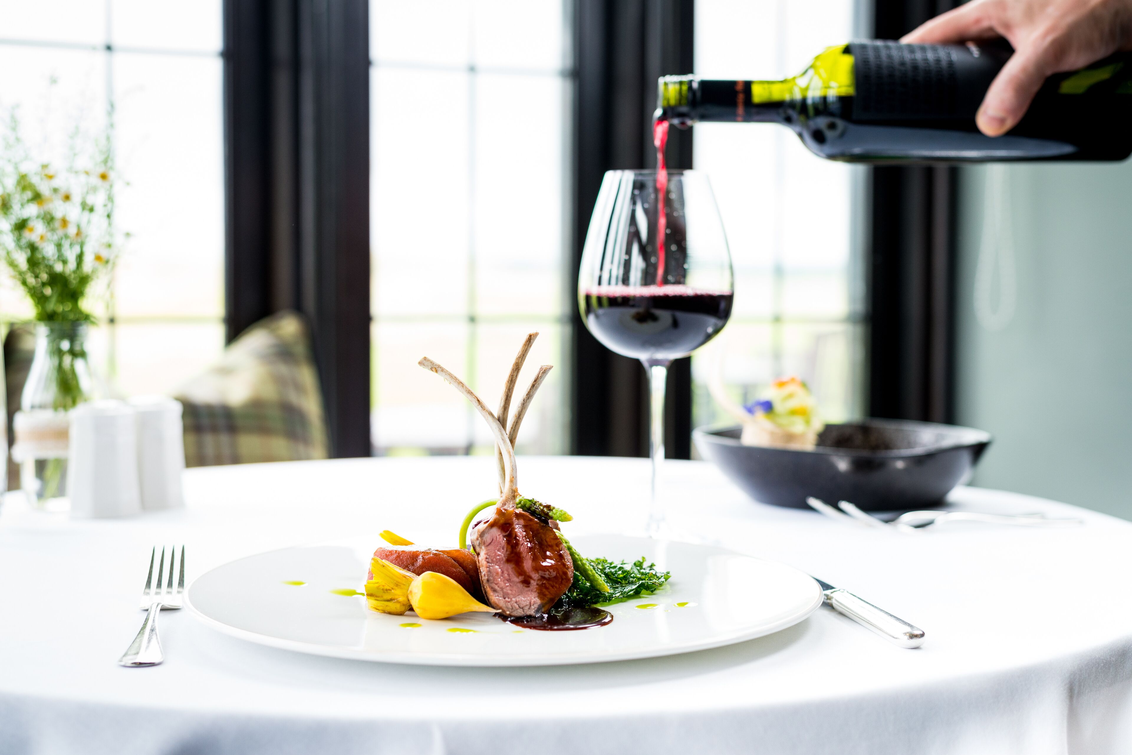 A dining table with a plate of food, a wine glass, and a bottle of wine being poured into the glass.