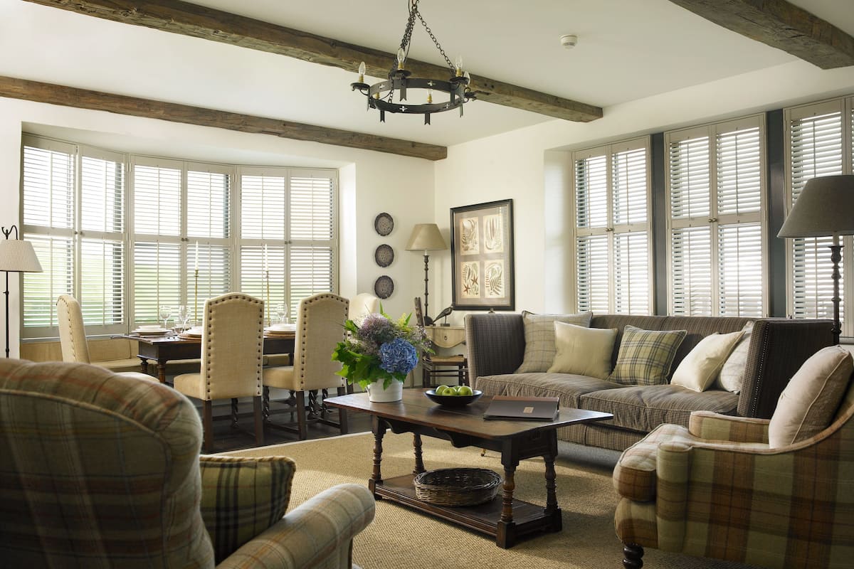 A spacious living room with a high ceiling, exposed wooden beams, a chandelier, and large windows with blinds.