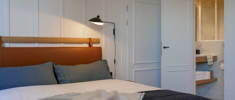 A neatly made bed with a wooden headboard, a bedside lamp, and a shelf with towels and toiletries.