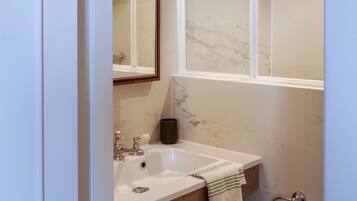 A bathroom with a marble wall, a sink with a towel rack, a mirror, and a shower with a ceiling light.