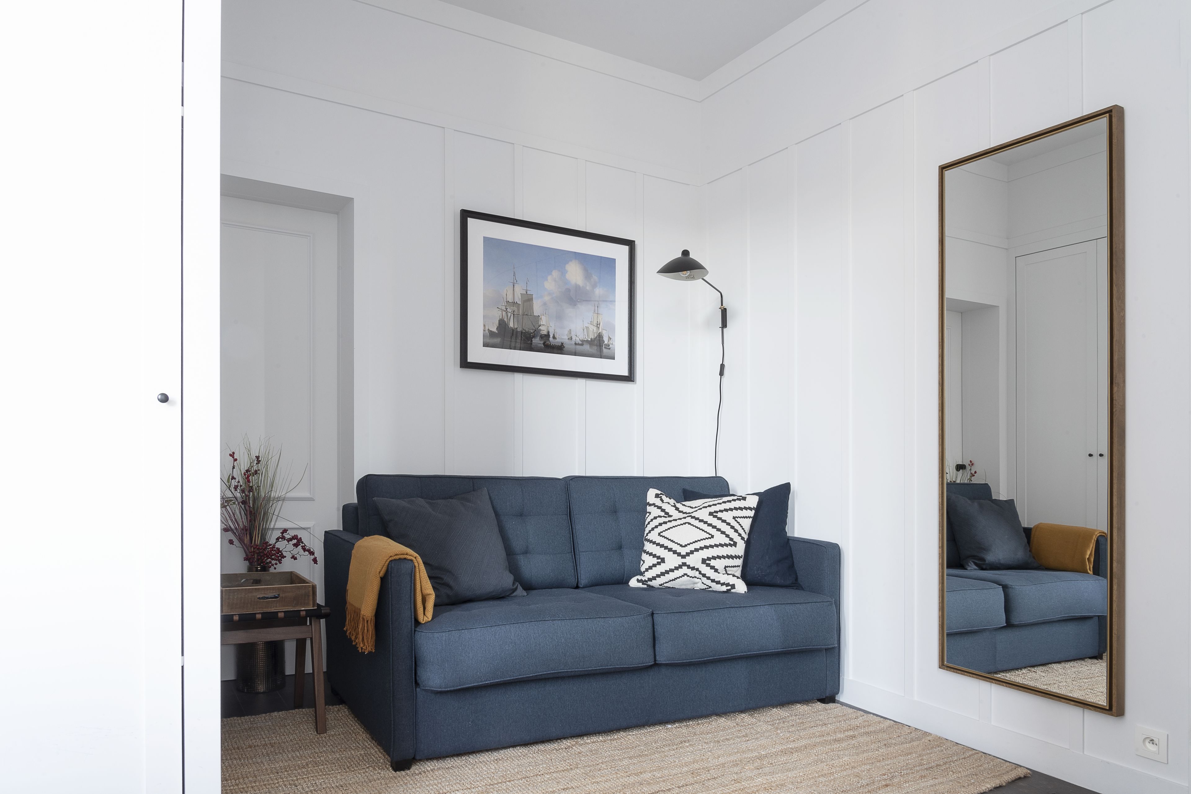 A modern living room with a dark blue sofa, a framed cityscape picture, a floor lamp, and a large mirror.