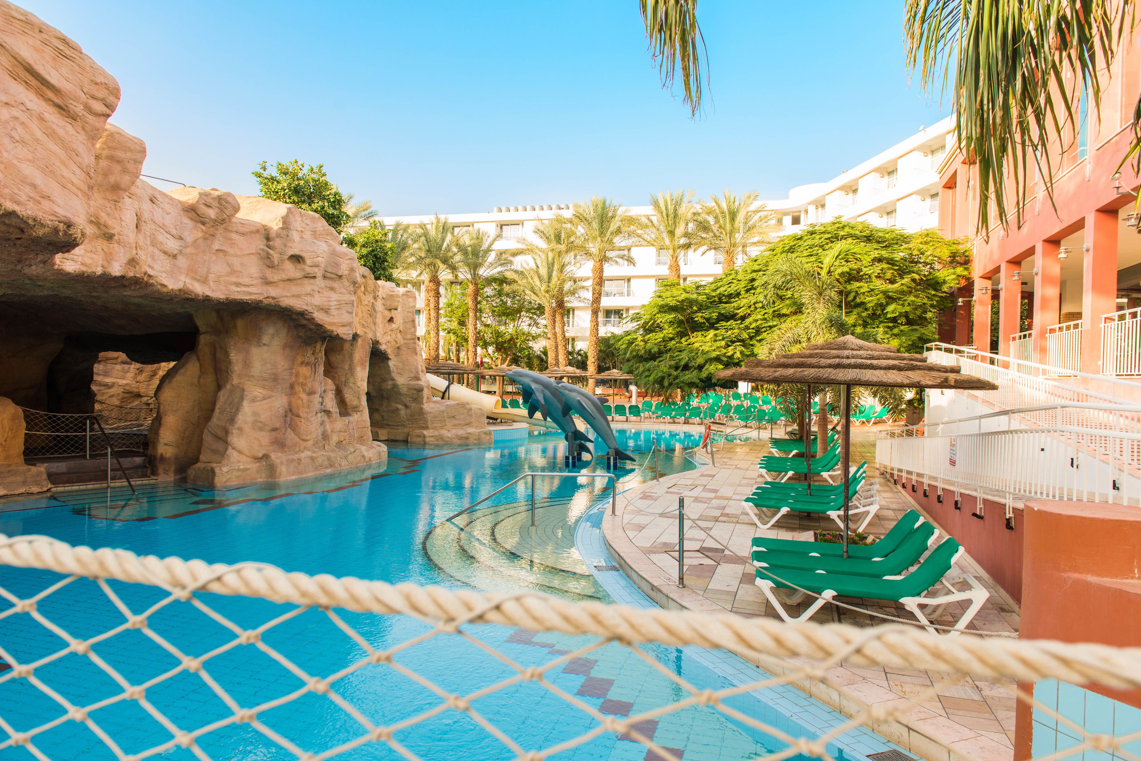 A swimming pool with a rock formation and a water slide, surrounded by lounge chairs and palm trees.