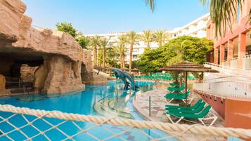 A swimming pool with a rock formation and a water slide, surrounded by lounge chairs and palm trees.