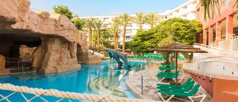 A swimming pool with a rock formation and a water slide, surrounded by lounge chairs and palm trees.