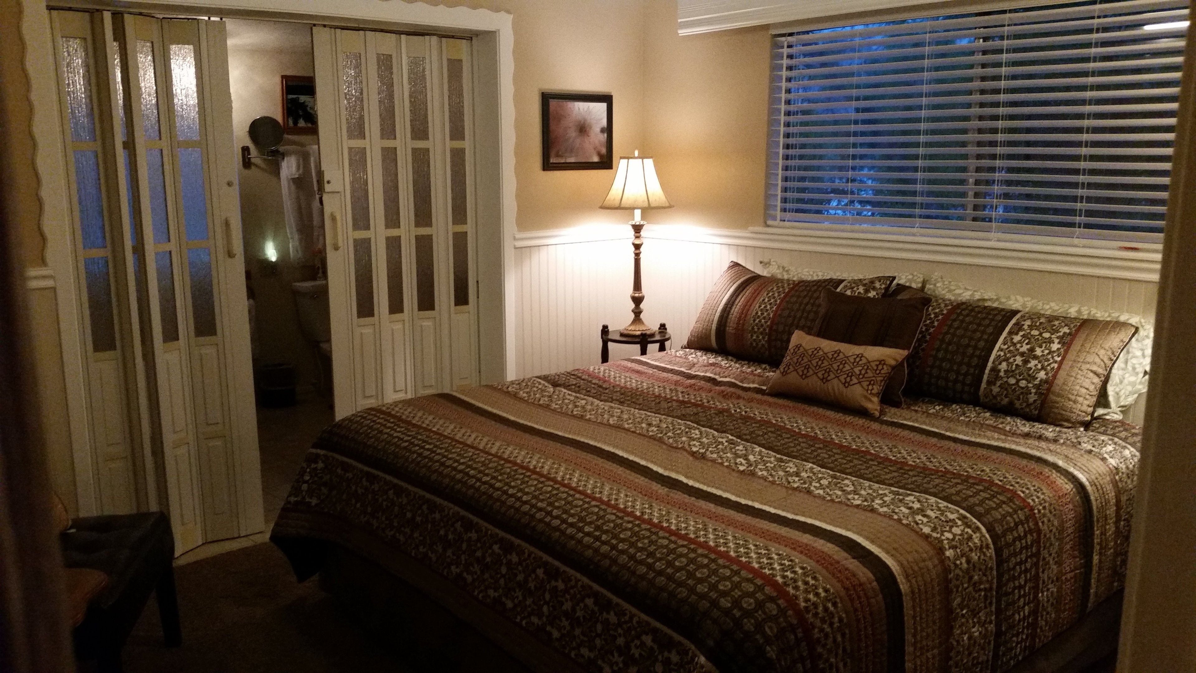 Quarto com cama, mesa de cabeceira, luminária, janela com persianas e porta para um banheiro.