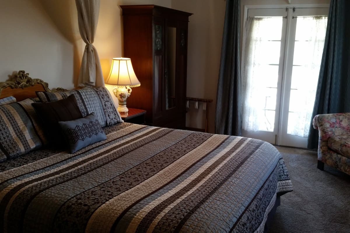 A bedroom with a large bed, a nightstand with a lamp, a wooden wardrobe, a floral sofa, and a door leading to a balcony.
