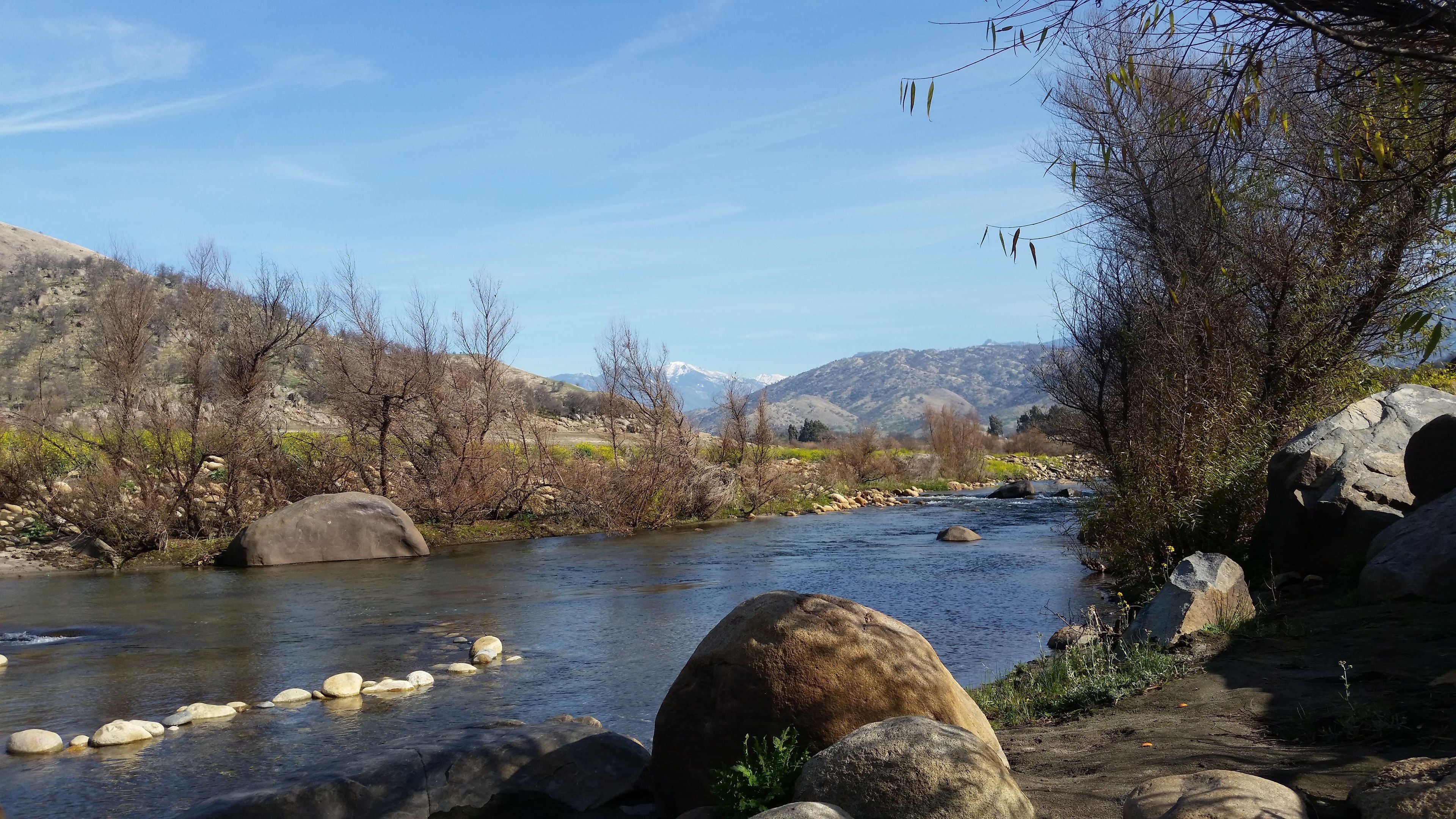A river with rocks and trees, surrounded by mountains and clear skies.