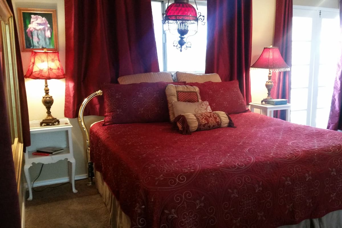 A neatly made bed with a red bedspread, two bedside tables with lamps, and a window with curtains.