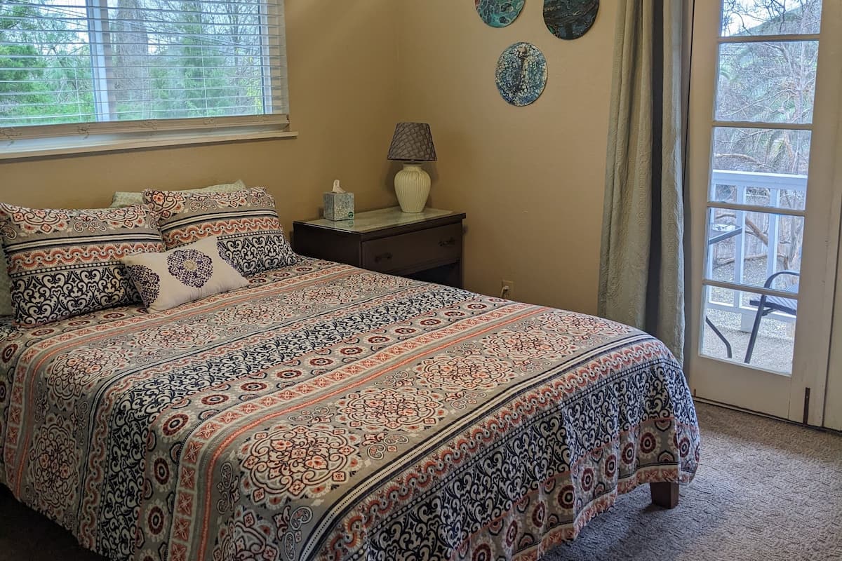 A bedroom with a bed, a ceiling fan, a nightstand with a lamp, a window with blinds, and a door with a view of the outside.