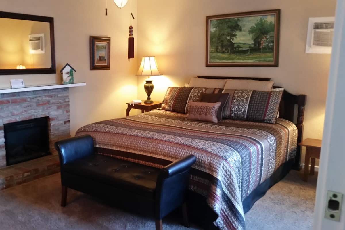 A bedroom with a fireplace, a bed with patterned bedding, a nightstand with a lamp, and a chair.