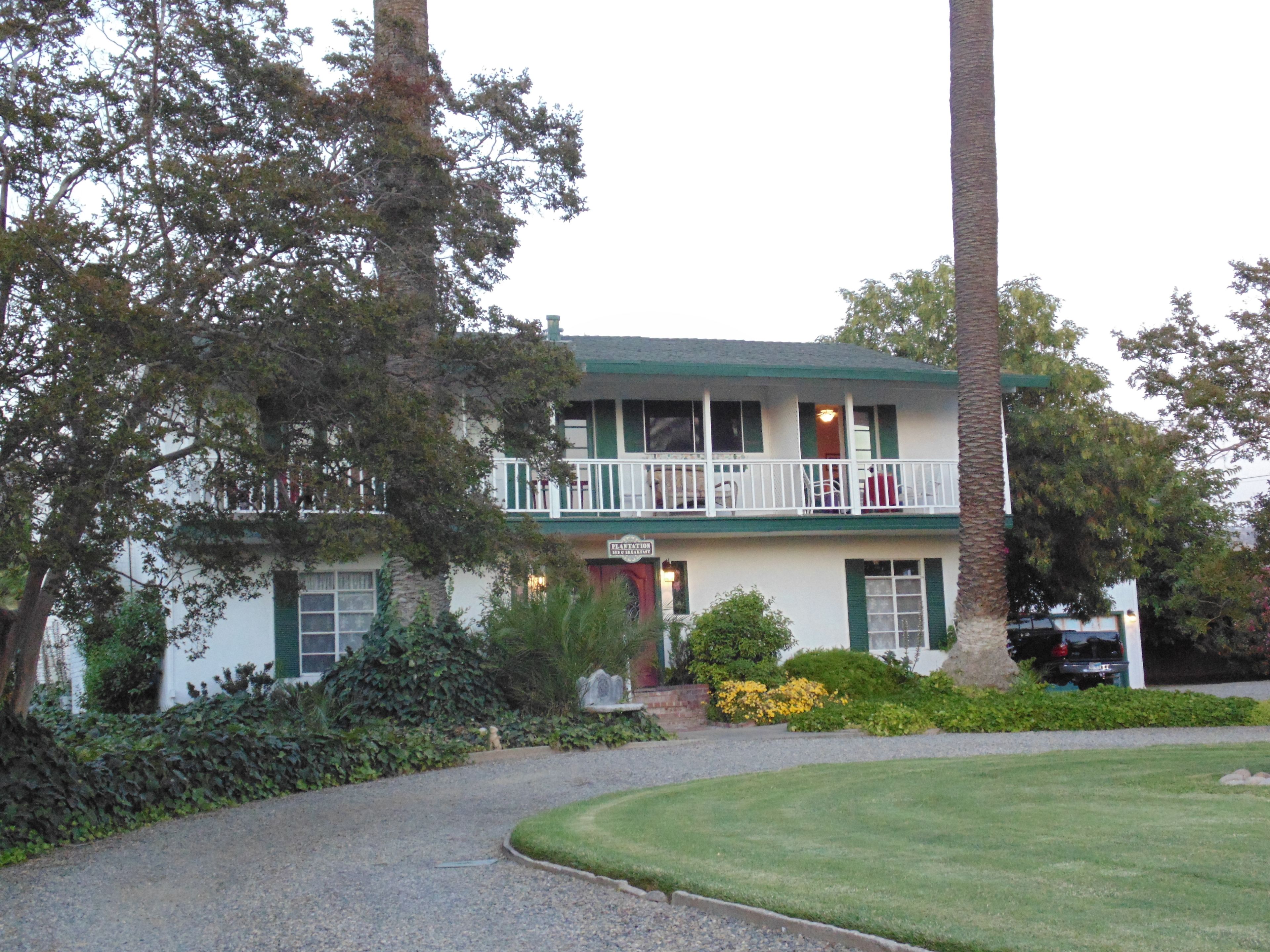 A two-story building with a balcony, surrounded by greenery and a gravel driveway.