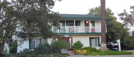 A two-story building with a balcony, surrounded by greenery and a gravel driveway.