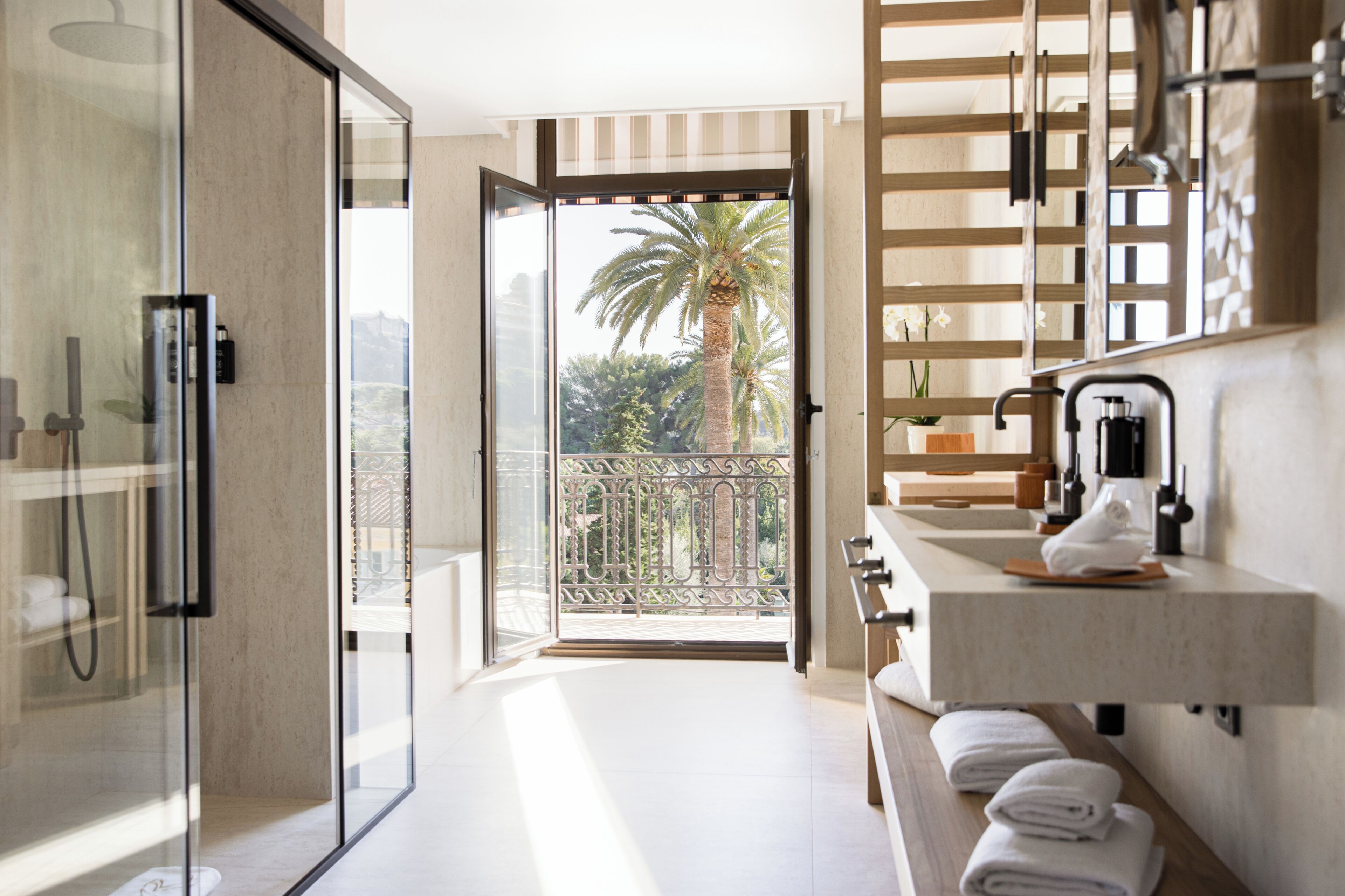 Casa de banho moderna com cabine de chuveiro em vidro, pia com porta-toalhas e vista para uma palmeira através de uma porta aberta.
