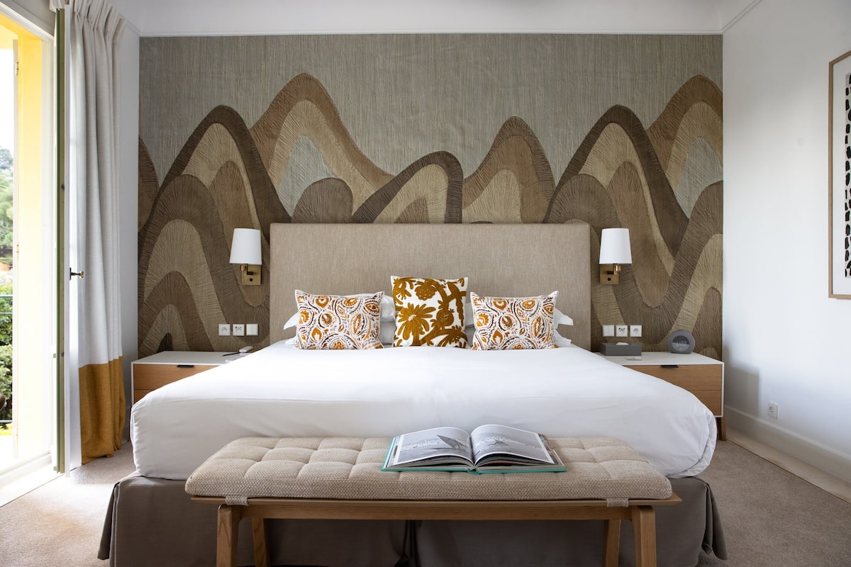 A spacious bedroom with a large bed, a bedside table with a lamp, and a bench with a book.