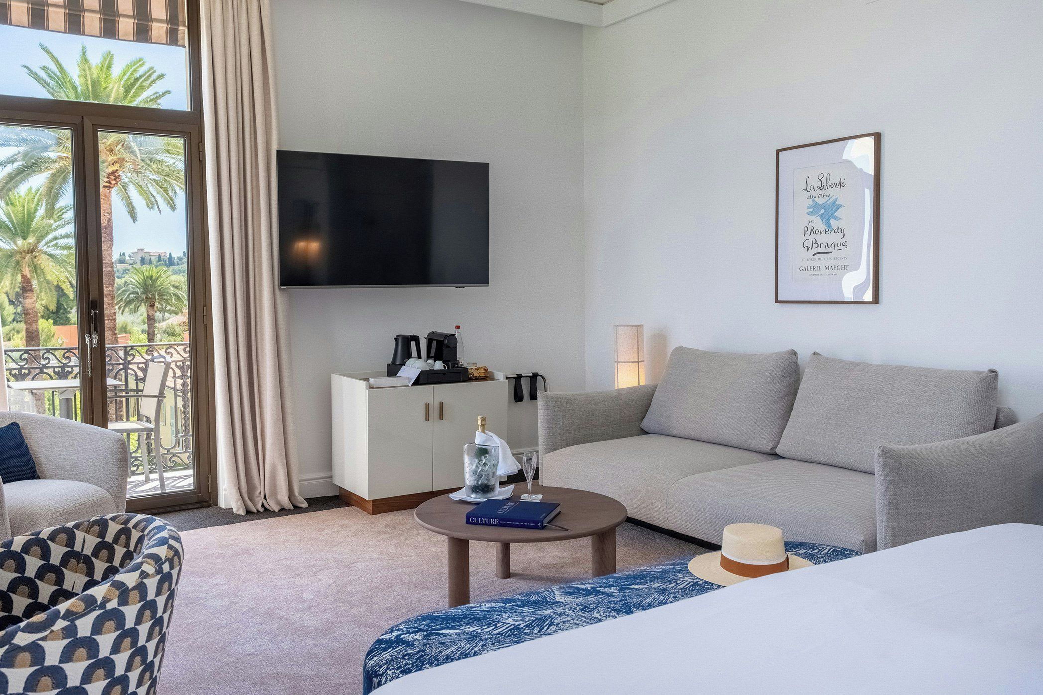 A hotel room with a sofa, a small table, a bed, a TV, and a balcony with palm trees.