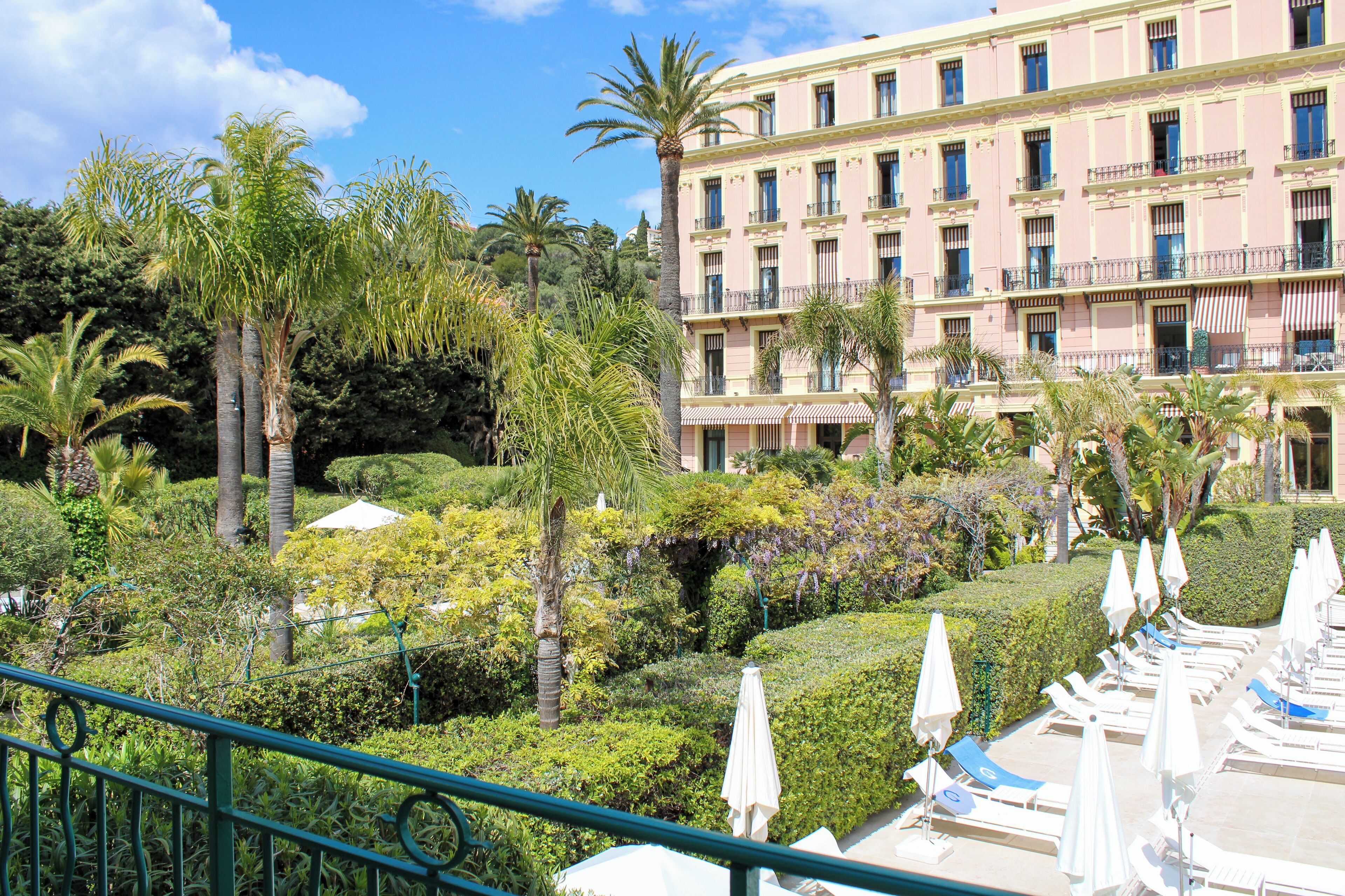 Un hotel con un jardín exuberante, palmeras y una zona de piscina con tumbonas y sombrillas.