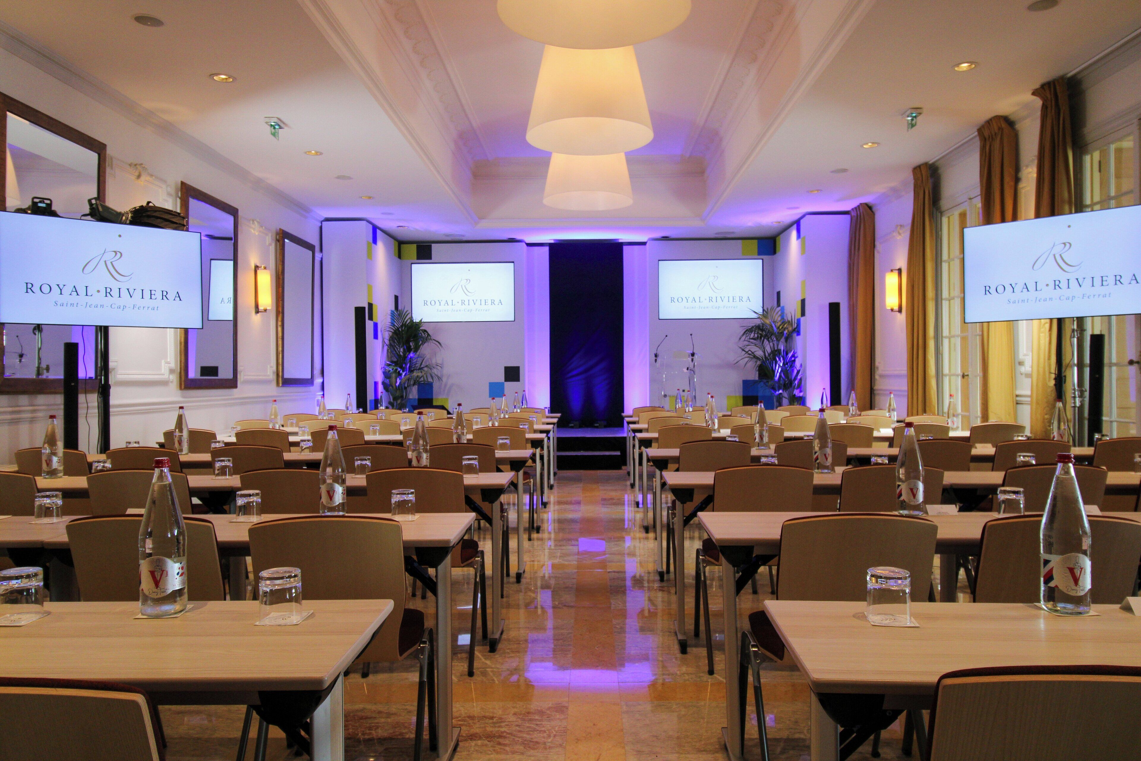 A conference room with tables, chairs, and presentation screens displaying the logo of Royal Riviera.