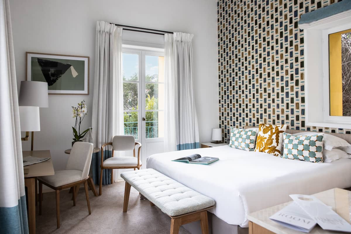 A hotel room with a bed, a desk, a chair, a bench, and a window with curtains.