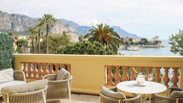 Un balcon doté de meubles en osier et d’une table, offrant une vue sur une zone côtière avec des montagnes et une étendue d’eau.