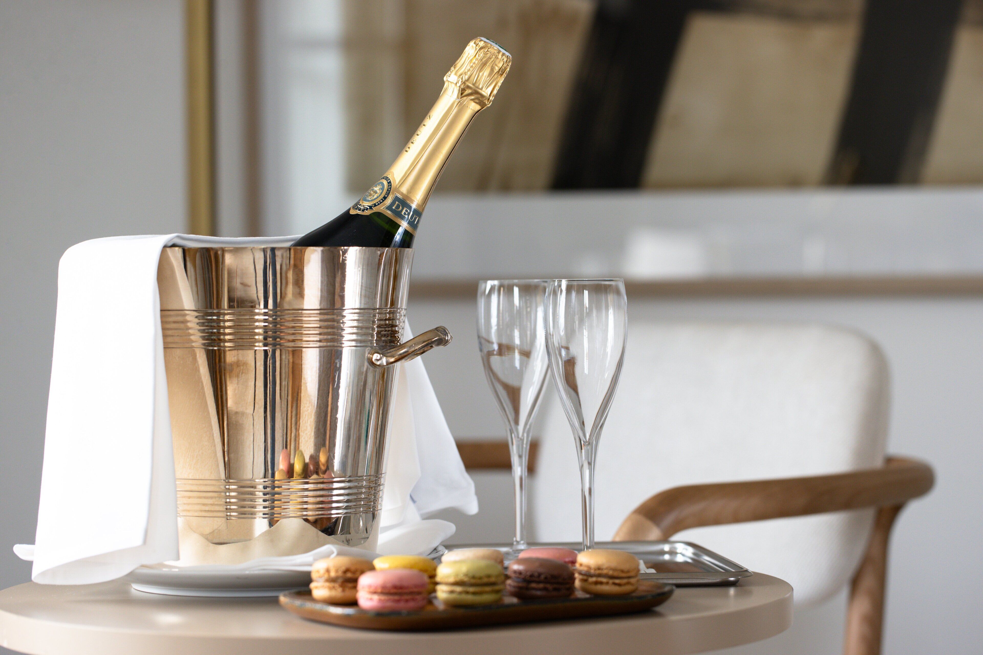 A champagne bottle in a metallic bucket, two empty wine glasses, a plate with macarons, and a knife on a round table.