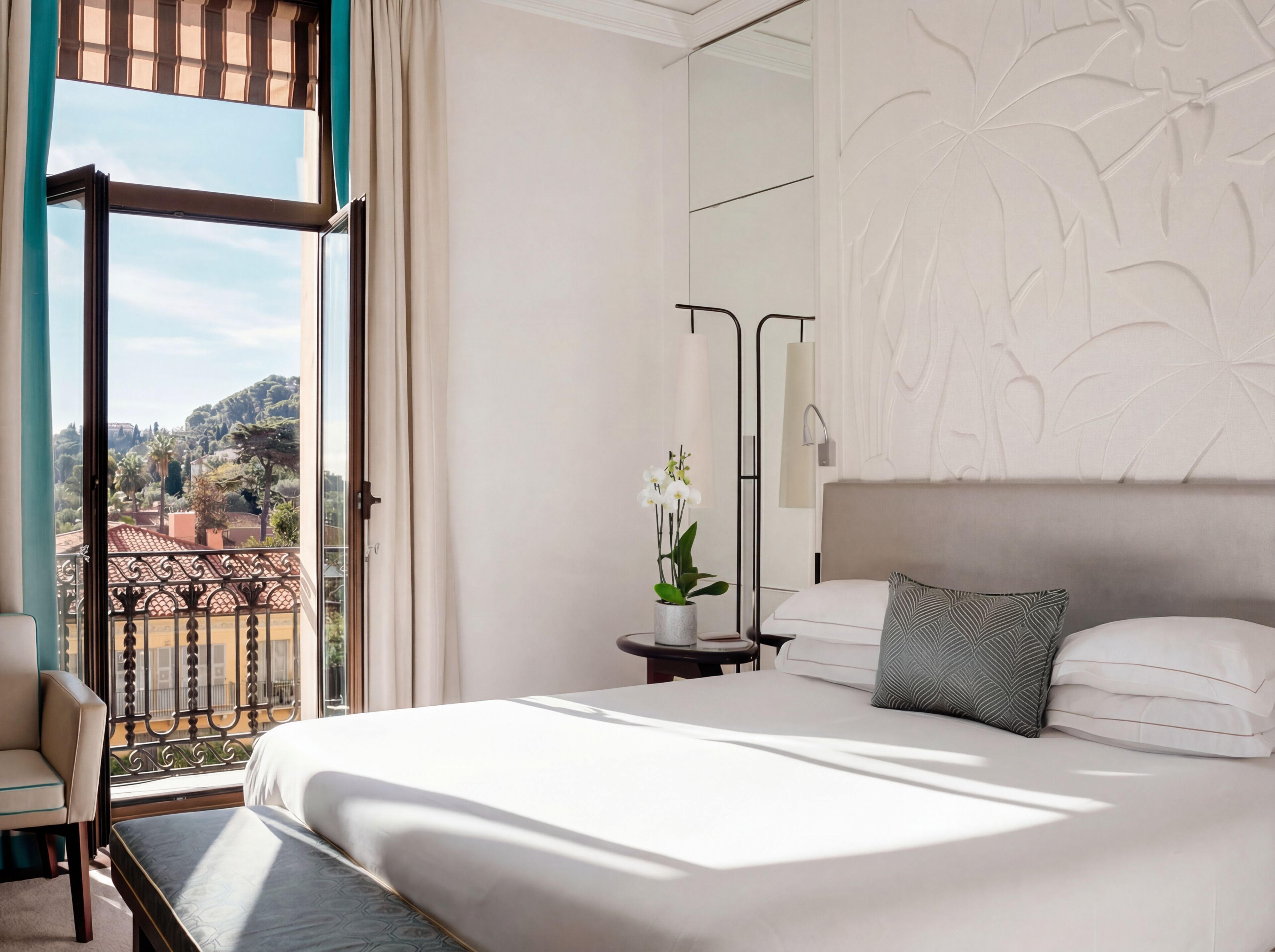 Une chambre d’hôtel comprenant un lit, une chaise, une petite table avec un vase de fleurs, une grande fenêtre donnant sur un paysage urbain et un panneau décoratif au mur.