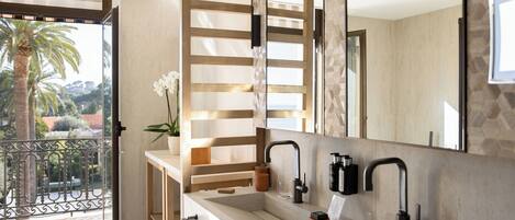 A modern bathroom with two sinks, a large mirror, and a view of a balcony with a palm tree.