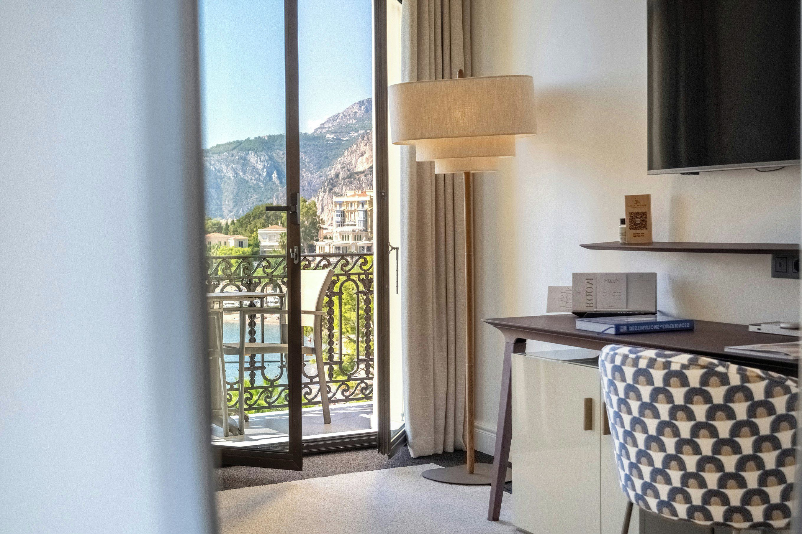 A hotel room with a balcony, a desk with a lamp, and a TV mounted on the wall.