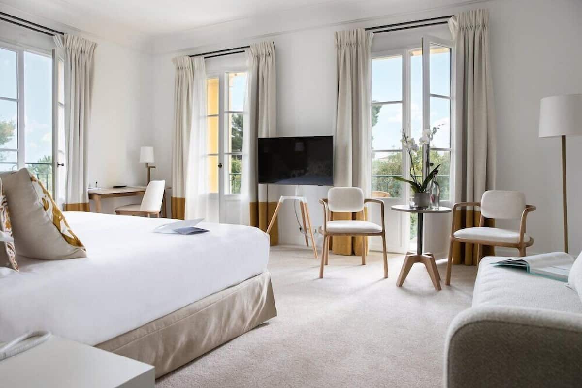 A hotel room with a bed, two chairs, a small table, a TV, and large windows with curtains.
